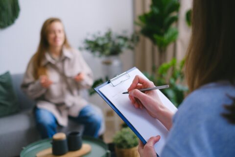 A person is writing on paper while another person sits in the background.